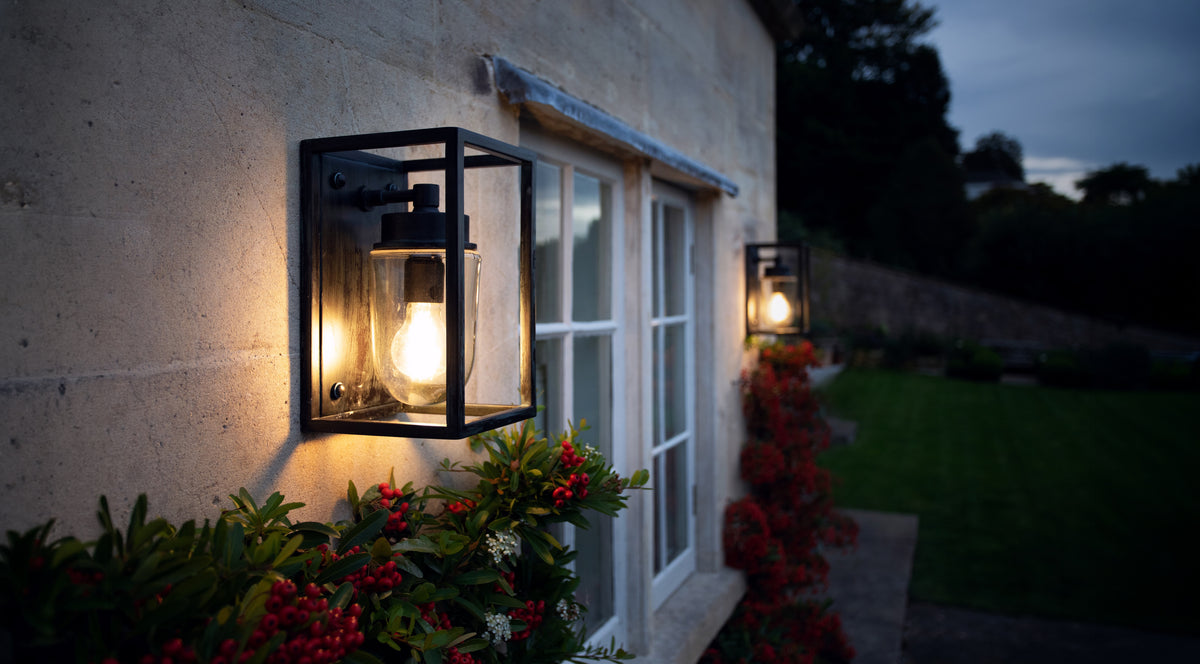 Two agriframes sherbourne outdoor open box lanterns installed on a stone house exterior either side of a window with plants growing around the lights.