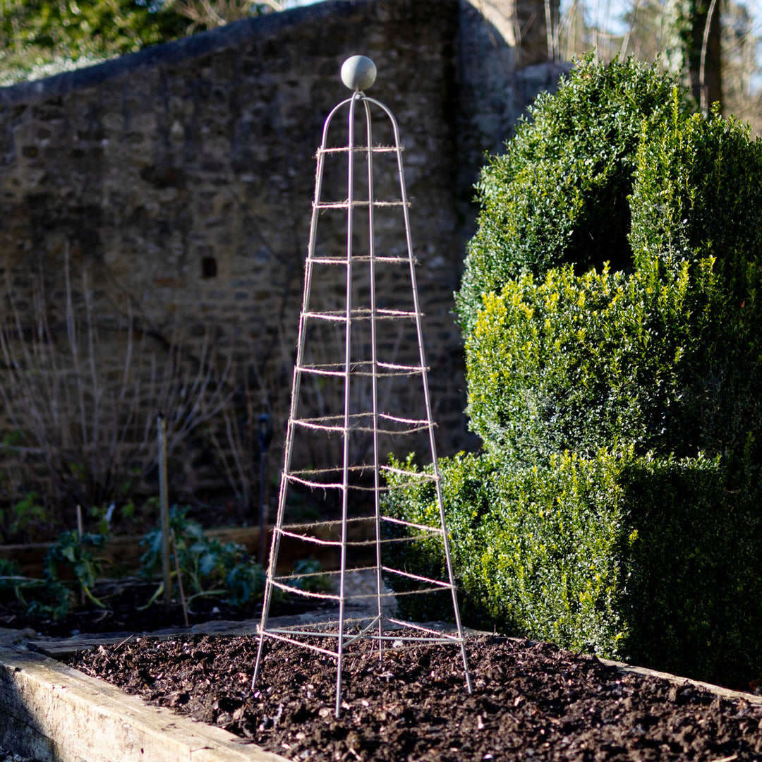Metal garden obelisk in a garden setting with a stone wall and bushes.