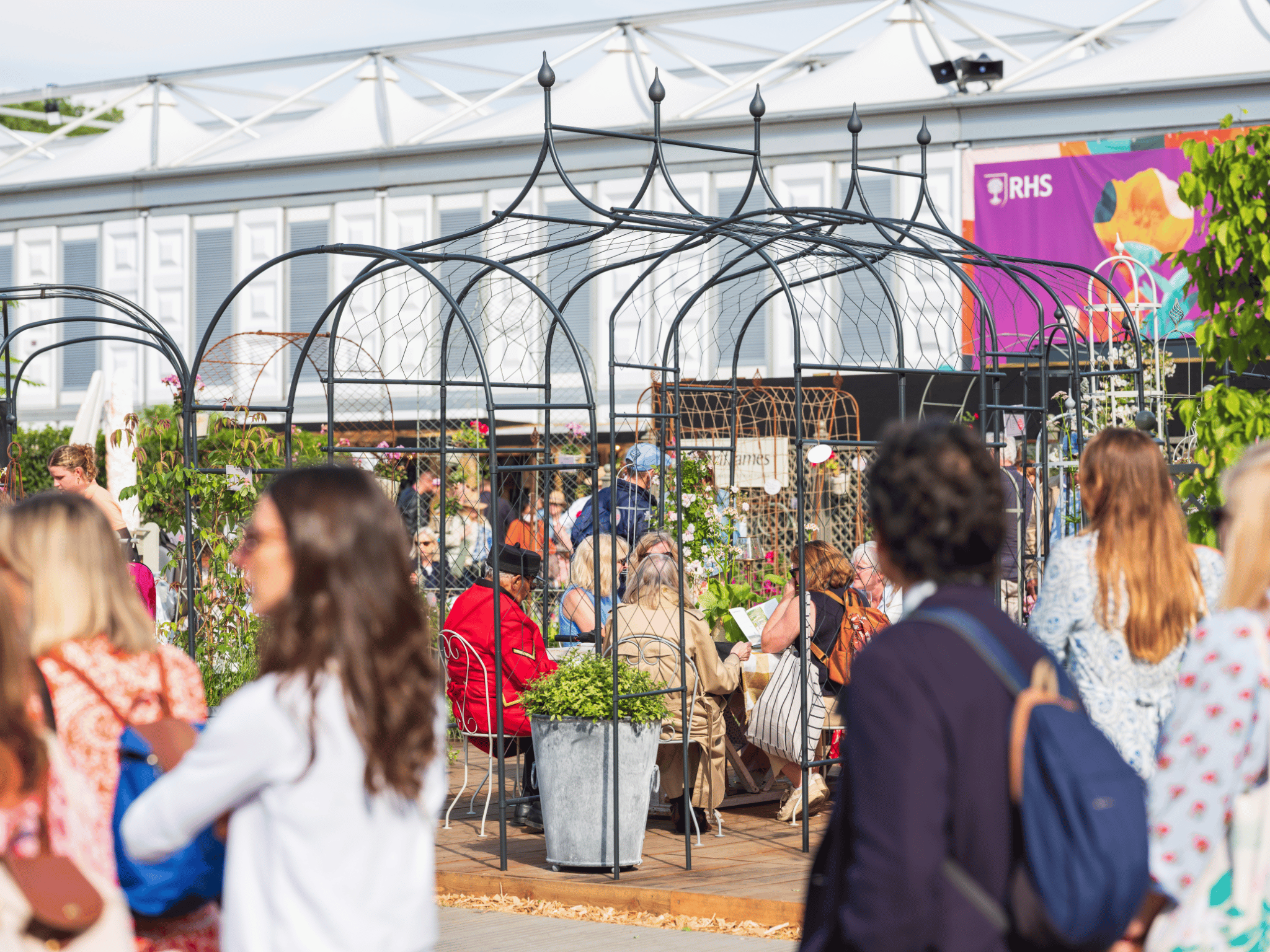People visiting a garden show with a metal archway and plants in the background.