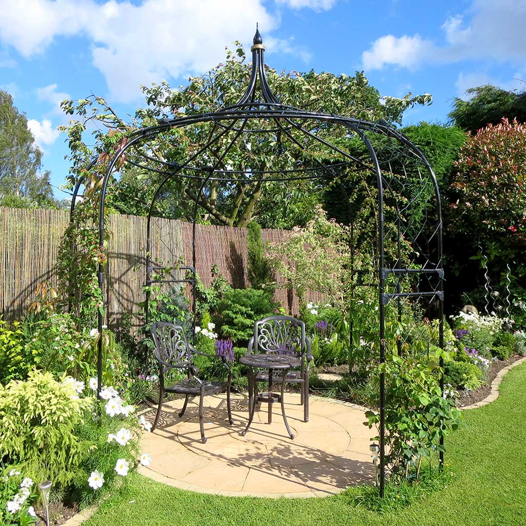 Garden setting with a black metal arbor, chairs, and a table surrounded by greenery and flowers.