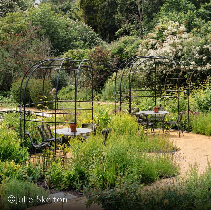 Two agriframes round arbours on a patio with tables and chairs underneath them and surrounded by flowerbeds