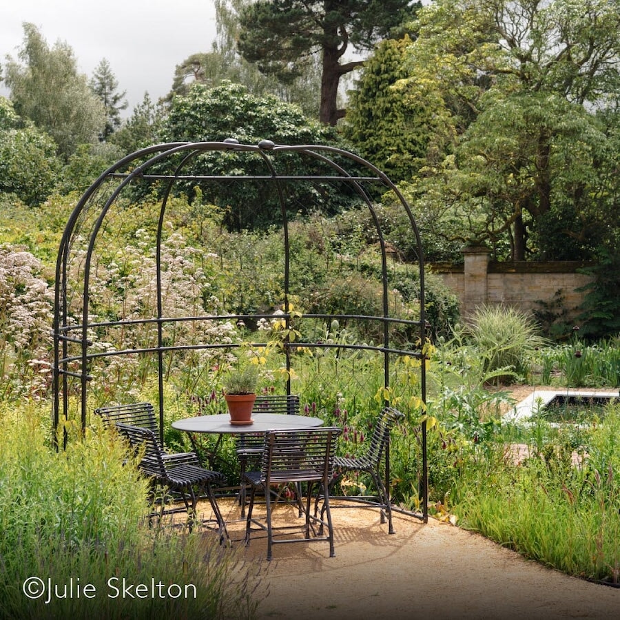 Agriframes Round arbour at the end of a garden path with a table and chair set underneath it