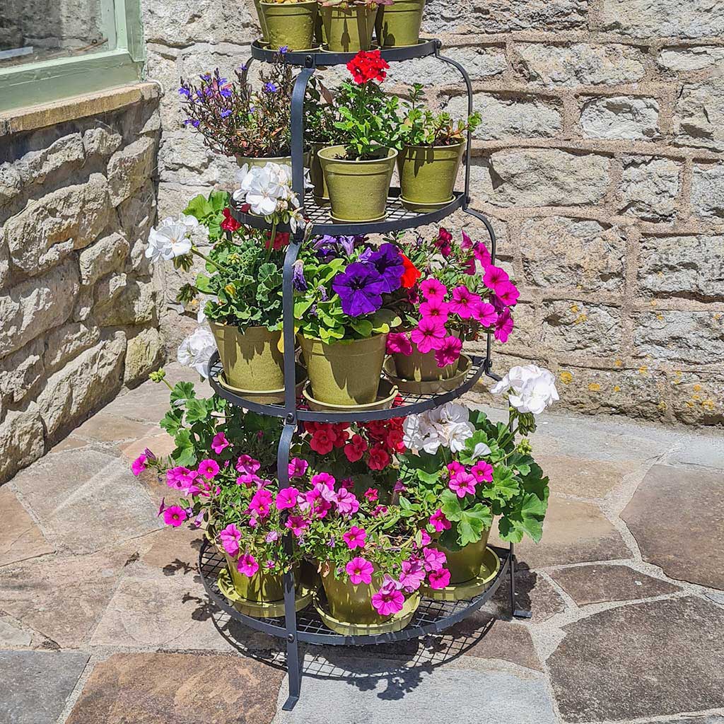 Three-tiered metal plant stand with potted flowers against a stone wall.