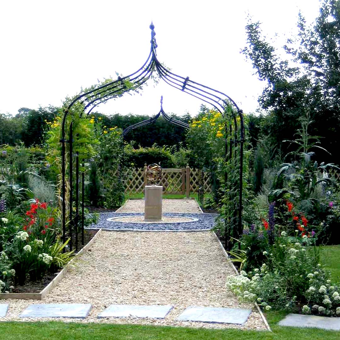 Gothic arch over gravel path, with garden sculpture and colourful flower beds
