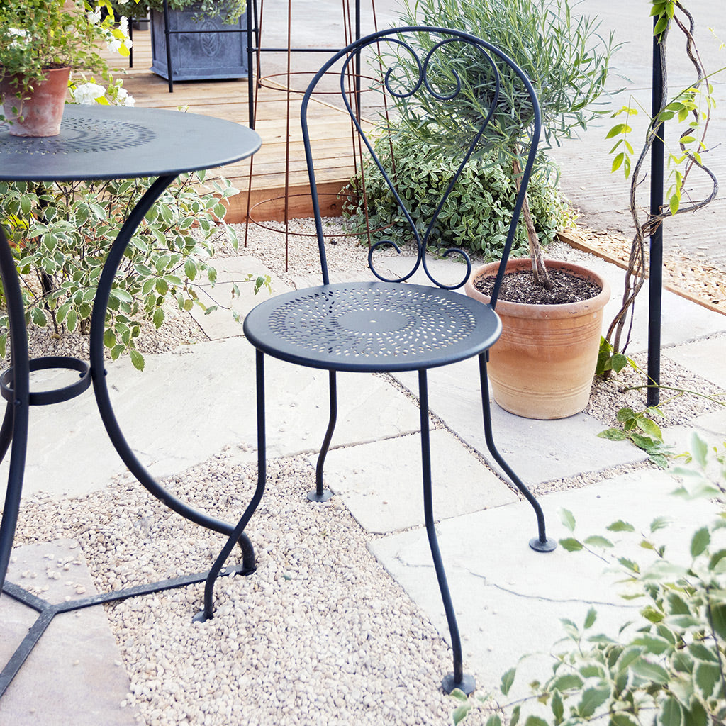 Black metal garden chair and table set in a garden setting with plants around.