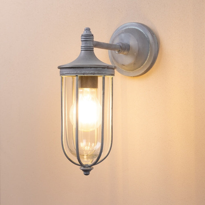 Agriframes hampstead outdoor wall lamp in an old iron finish with its bulb lit on a neutral wall. 