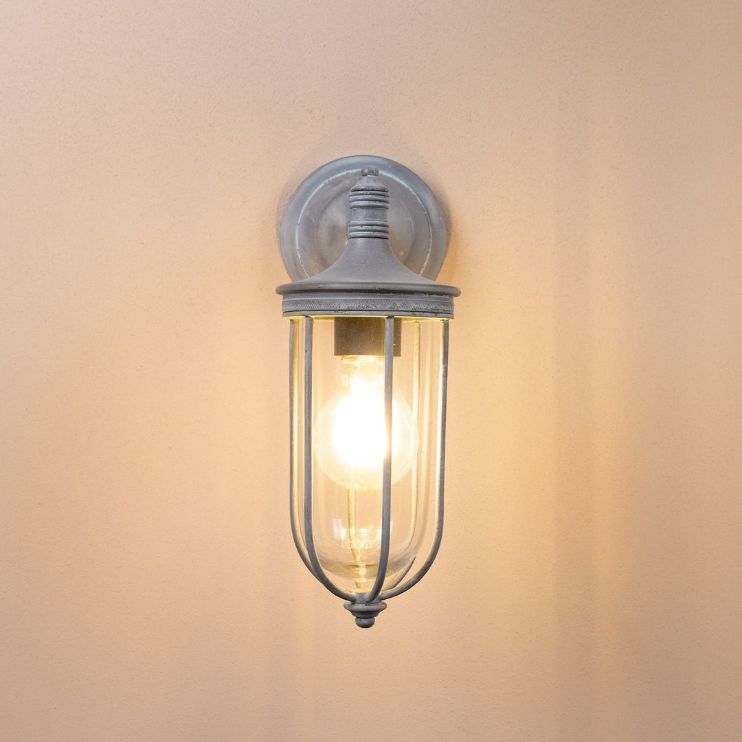 Agriframes hampstead outdoor wall lamp in an old iron finish with its bulb lit on a neutral wall. 