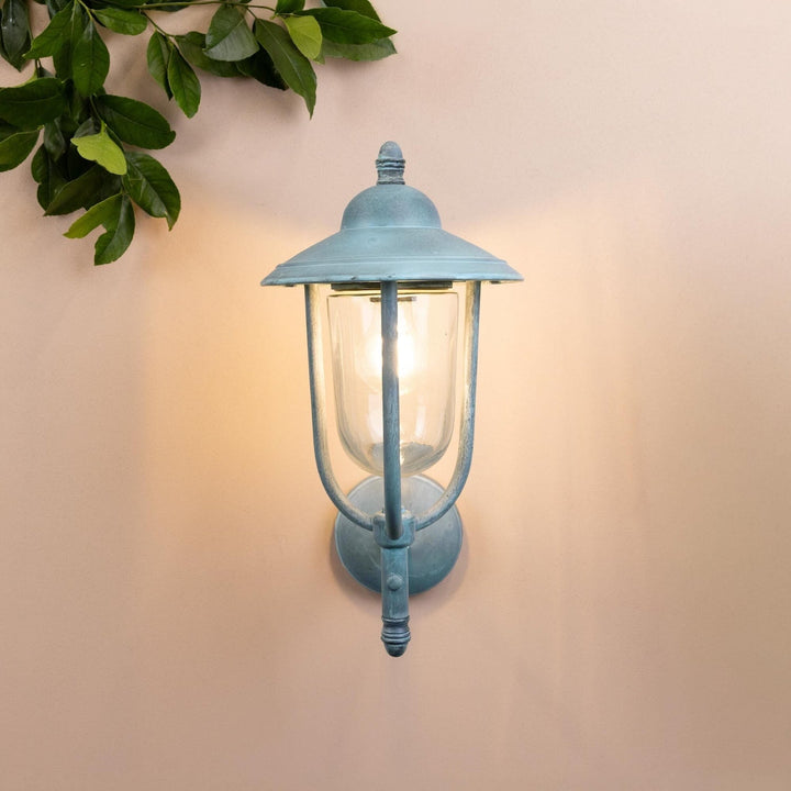 Agriframes kensington outdoor wall lamp in a verdigris finish with its bulb lit on a neutral coloured wall with decorative leaves. 