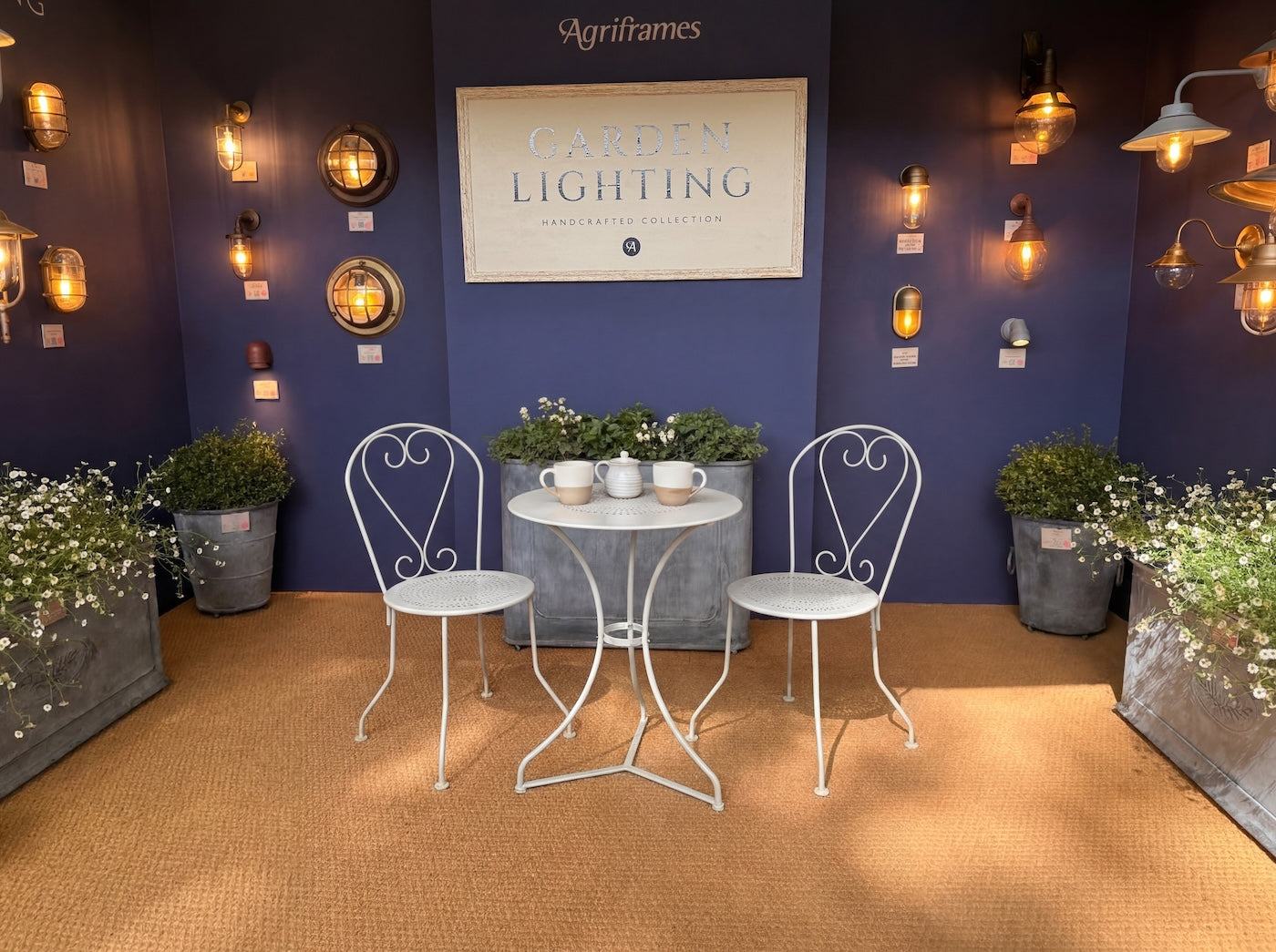 Agriframes' Outdoor Lighting display at RHS Chelsea Flower Show 2026, with various light fixtures on a blue wall and a chair and table set in the the middle of the room
