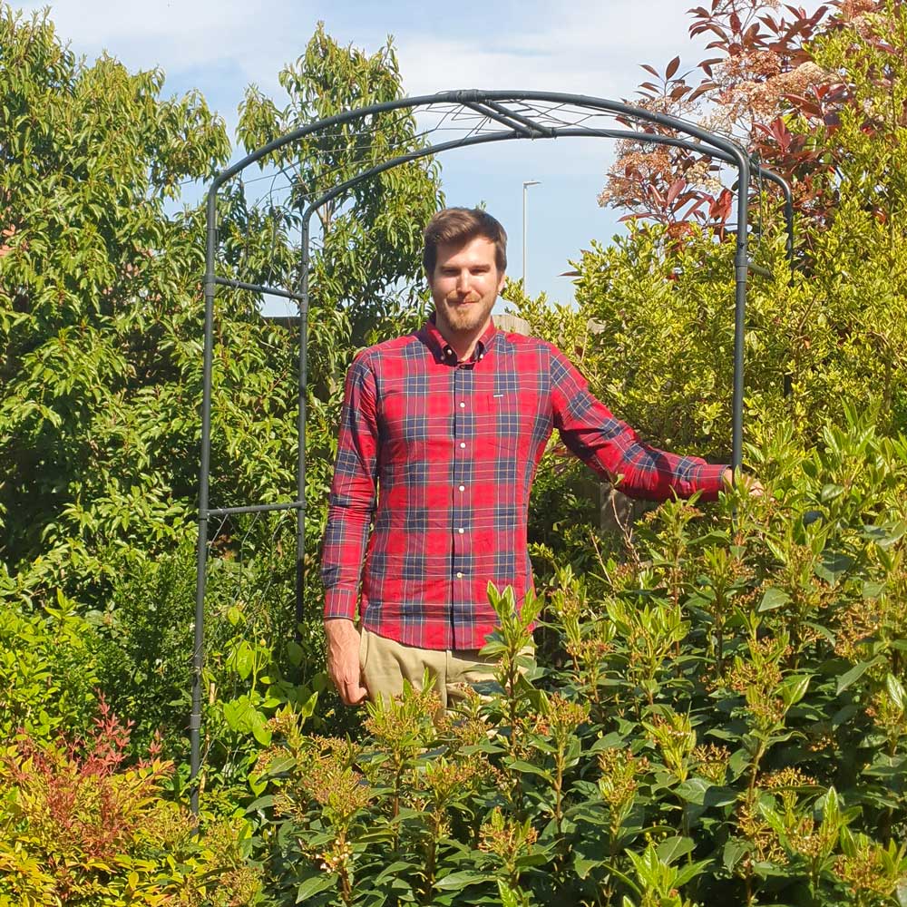 A man wearing a red plaid shirt and beige chinos stands underneath the Agriframes matte black Monet arch amongst a verdant garden scene. 