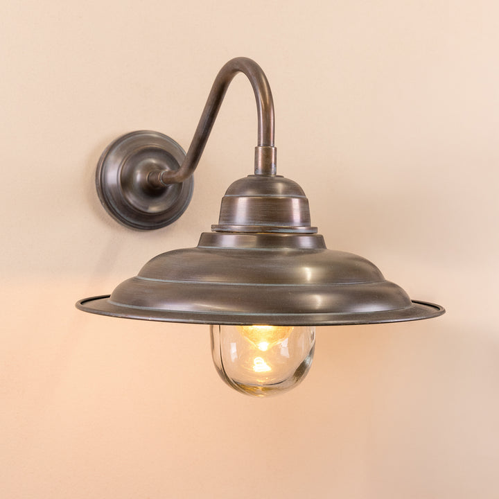 Angled image of the Agriframes Bucklebury Outdoor Wall Lamp in a Copper Patina finish with an illuminated bulb, mounted on a neutral background. 