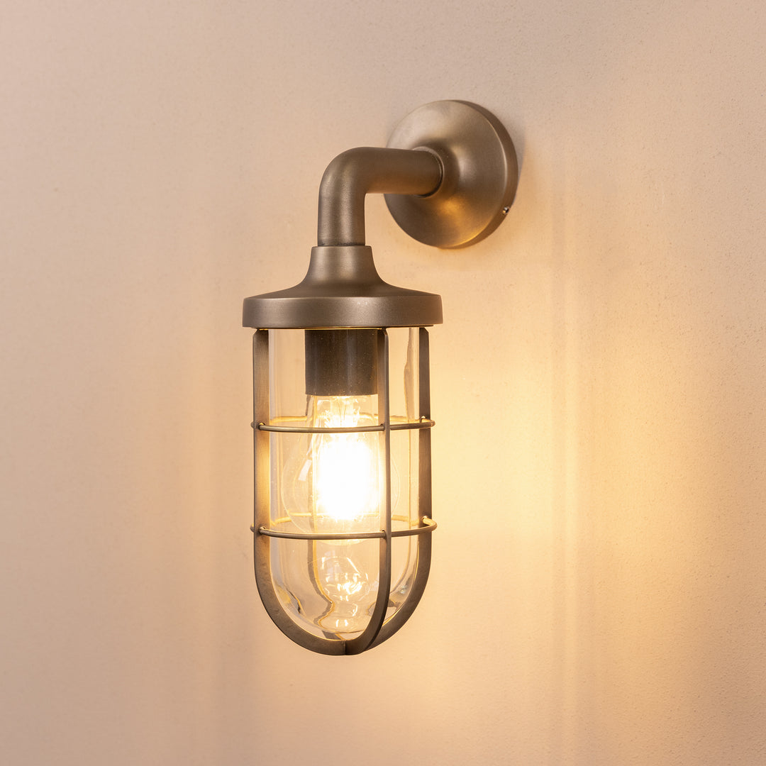 Angled image of the Agriframes Exmouth Outdoor Wall Lamp in a Brass finish with its bulb illuminated, mounted on a neutral background. 