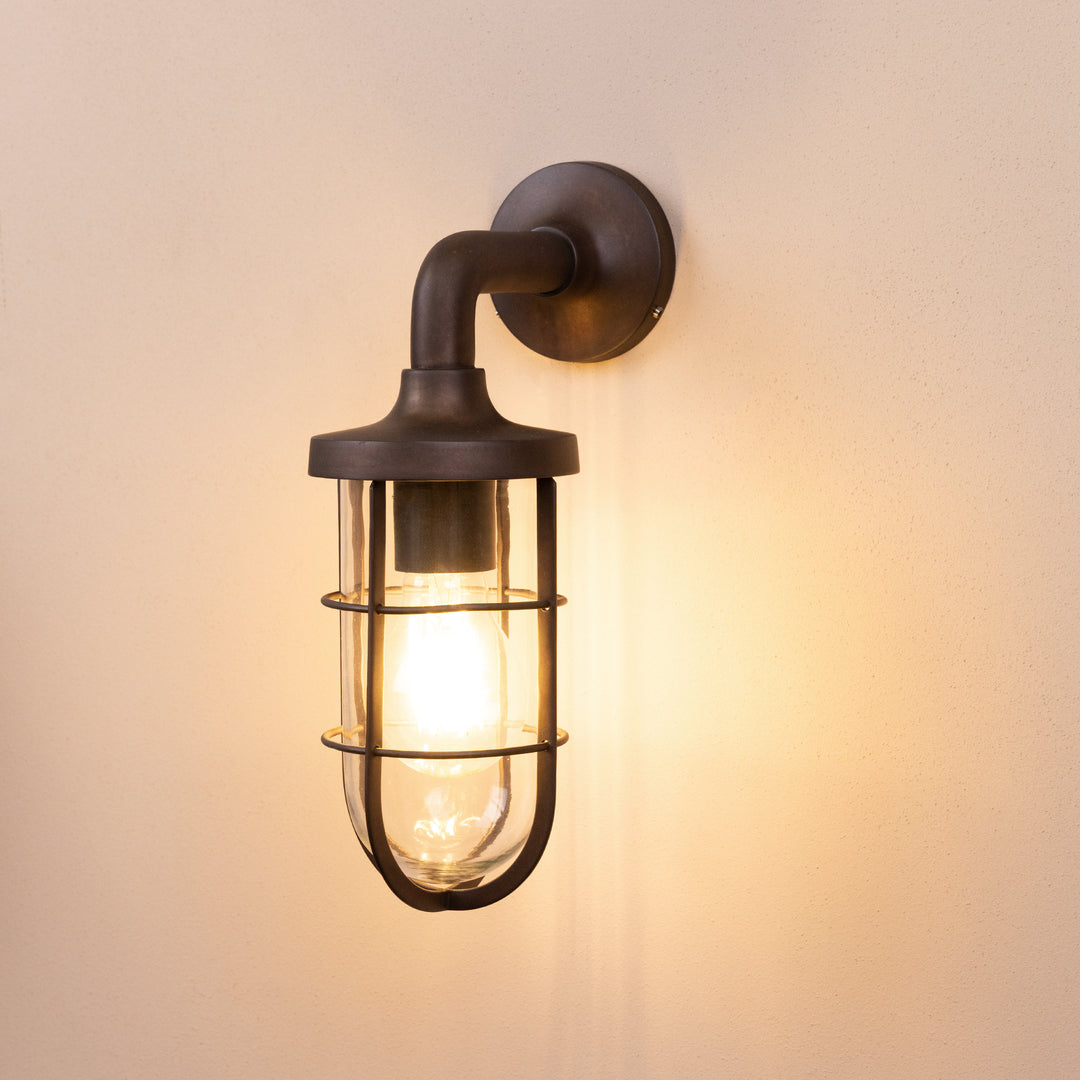 Angled image of the Agriframes Exmouth Outdoor Wall Lamp in a Bronze finish with its bulb illuminated, mounted on a neutral background. 