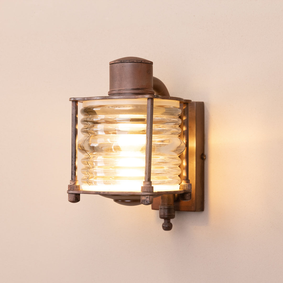 Angled image of the Agriframes From Outdoor Wall Lamp in a Rust finish with its bulb illuminated, mounted on a neutral-coloured wall. 