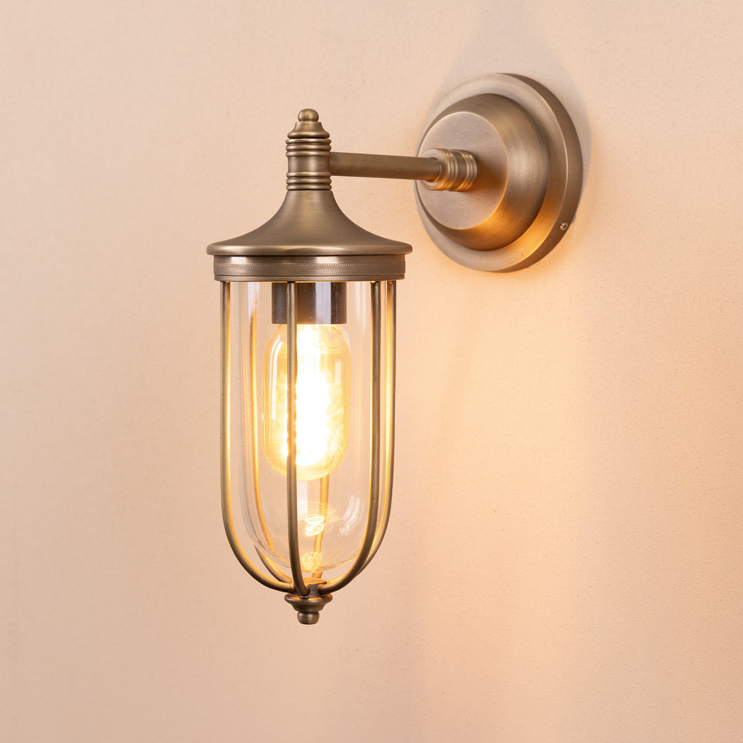 Angled image of the Agriframes Outdoor Wall Lamp in a Brass finish with its bulb illuminated, mounted on a neutral background. 
