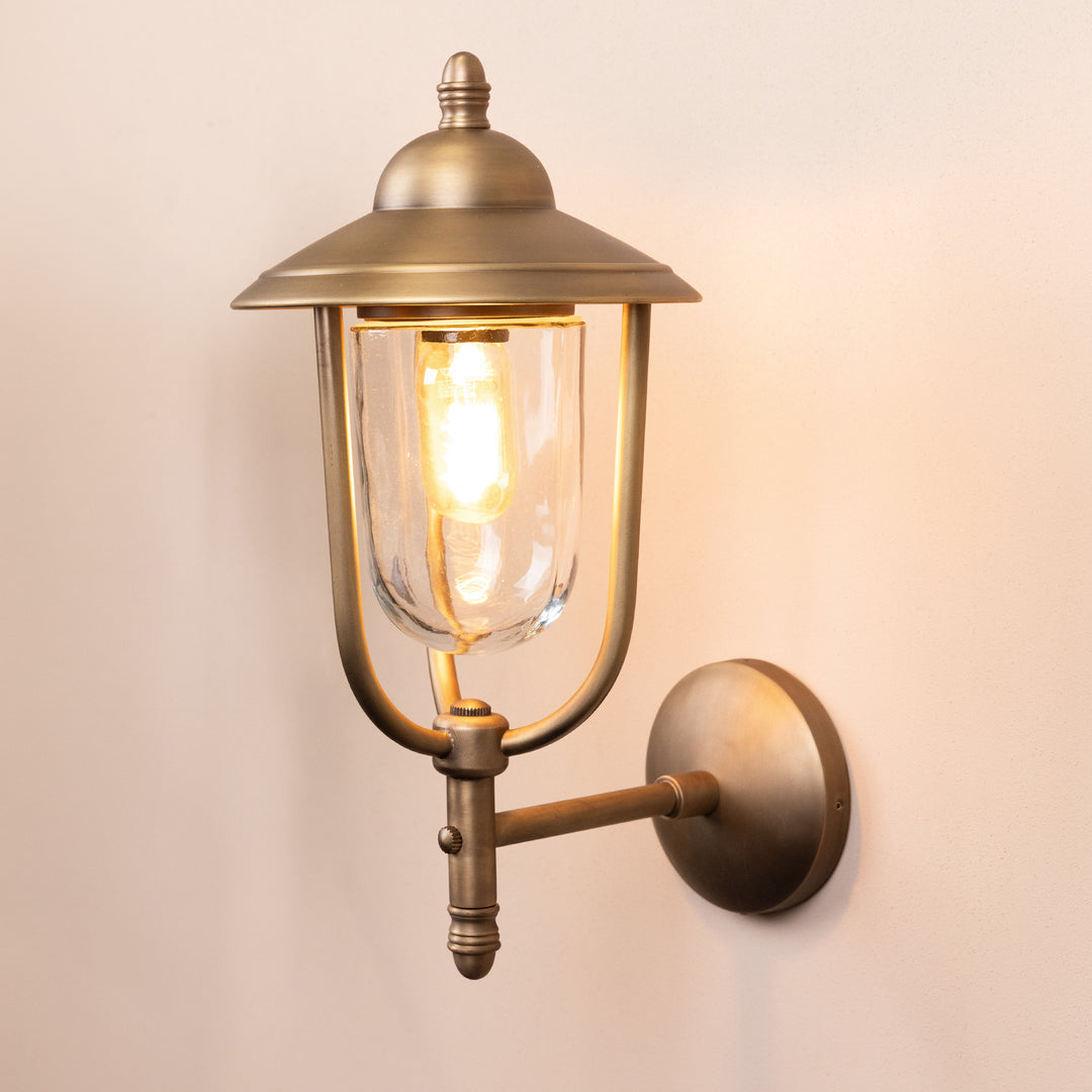 Angled image of the Kensington Outdoor Wall Lamp in a Brass finish with its bulb illuminated, mounted on a neutral background. 