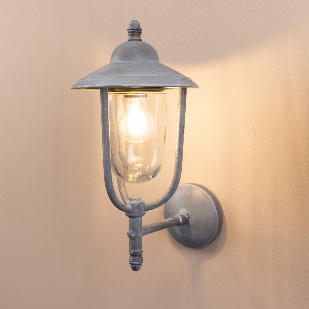 Angled image of the Agriframes Kensington Outdoor Wall Lamp in an Old Iron finish with its bulb illuminated, mounted on a neutral background. 