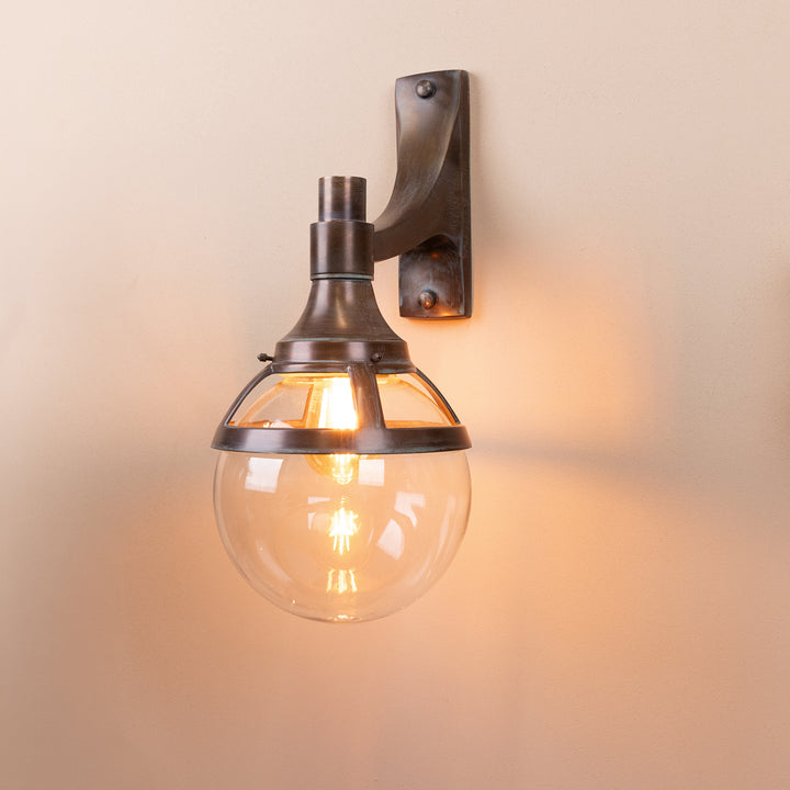 Angled image of the London Outdoor Wall Lamp in a Copper Patina finish with its bulb illuminated, mounted on a neutral background. 