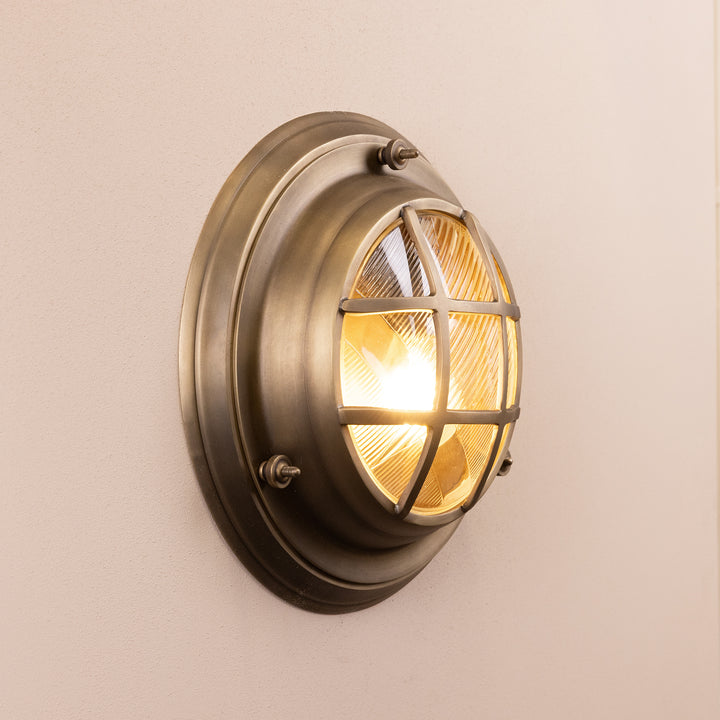 Angled image of the Agriframes Looe Bulk Head Round Lantern in a Brass finish with its bulb illuminated, mounted on a neutral background. 