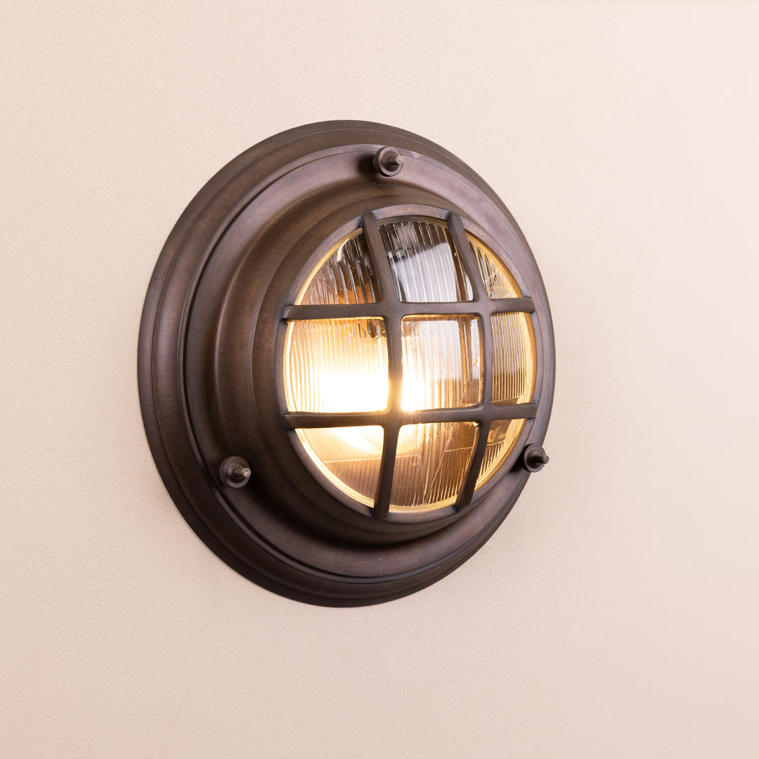Angled image of the Agriframes Looe Bulk Head Round Lantern in a Bronze finish with its bulb turned on, mounted on a neutral background. 
