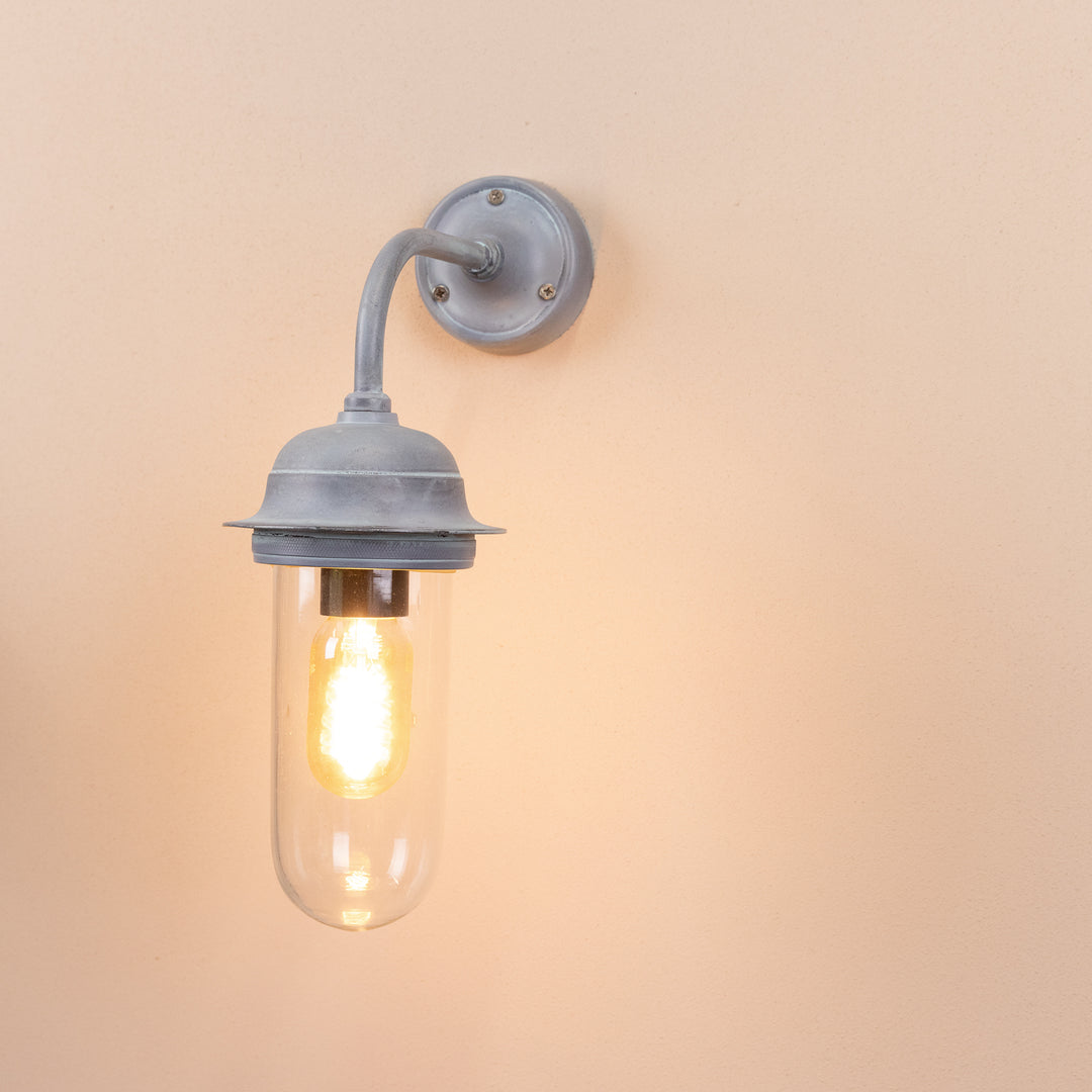 Angled image of the Agriframes Pewsey Outdoor Wall Lamp in an Old Iron finish with its bulb illuminated, mounted on a neutral background. 