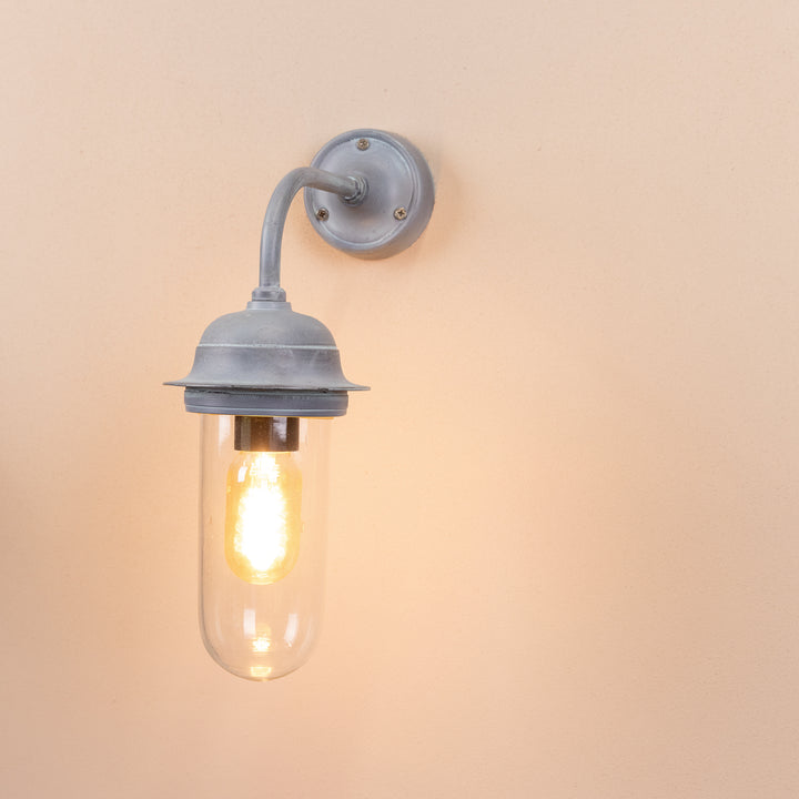 Angled image of the Agriframes Pewsey Outdoor Wall Lamp in an Old Iron finish with its bulb illuminated, mounted on a neutral background. 