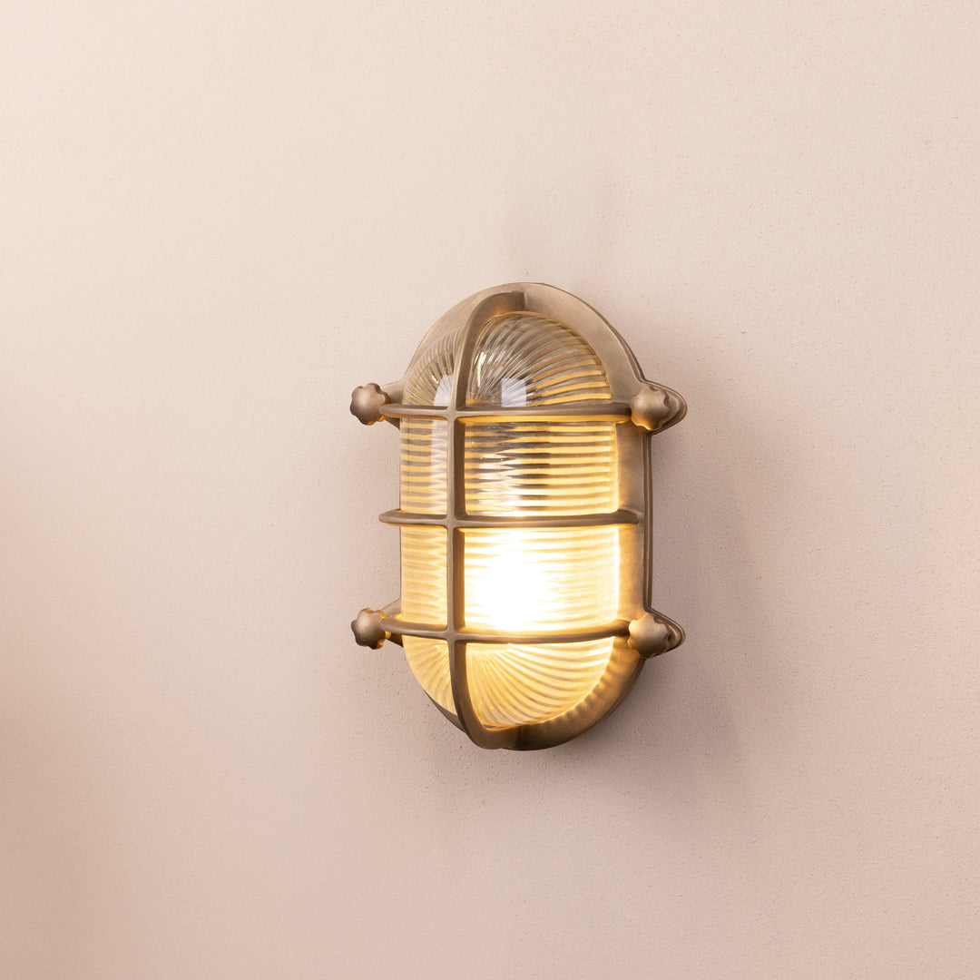 Angled mage of the Agriframes Bulk Head Oval Lantern in a Brass finish with its bulb illuminated, mounted on a neutral background. 