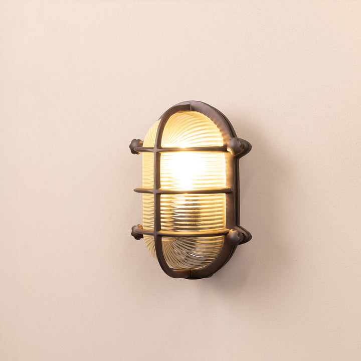 Angled image of the Agriframes Salcombe Bulk Head Oval Lantern in a Bronze finish with its bulb illuminated, mounted on a neutral background. 