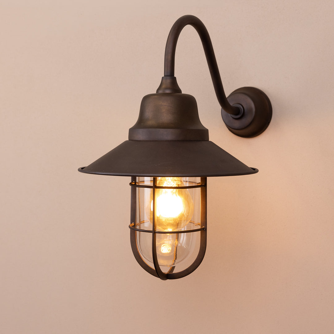 Angled image of the Agriframes Sidmouth Outdoor Wall Lamp in a Bronze finish with its bulb illuminated, mounted on a neutral background. 