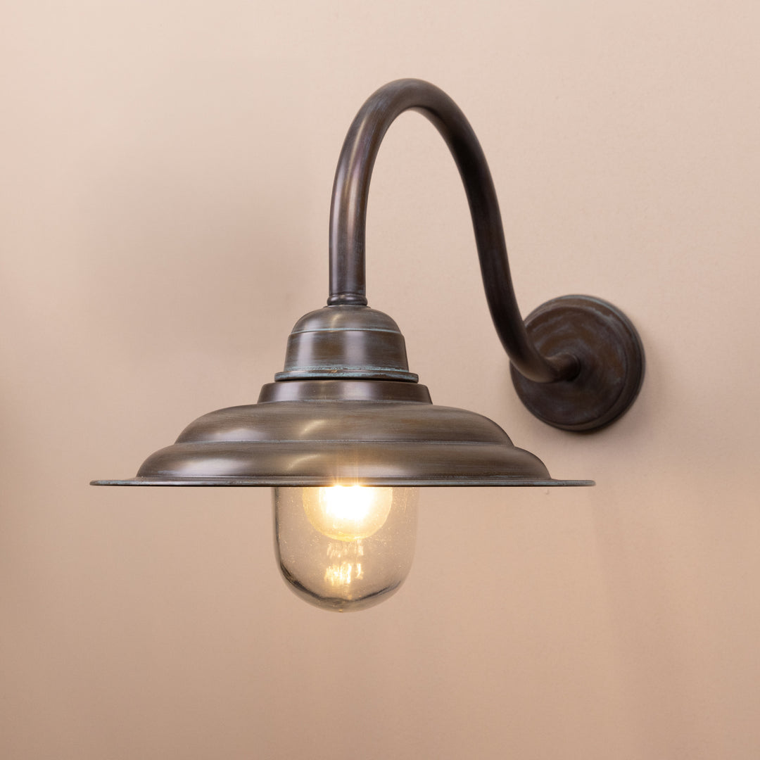 Angled image of the Agriframes Tetbury Outdoor Wall Lamp in a Copper Patina finish with its bulb illuminated, mounted on a neutral background. 