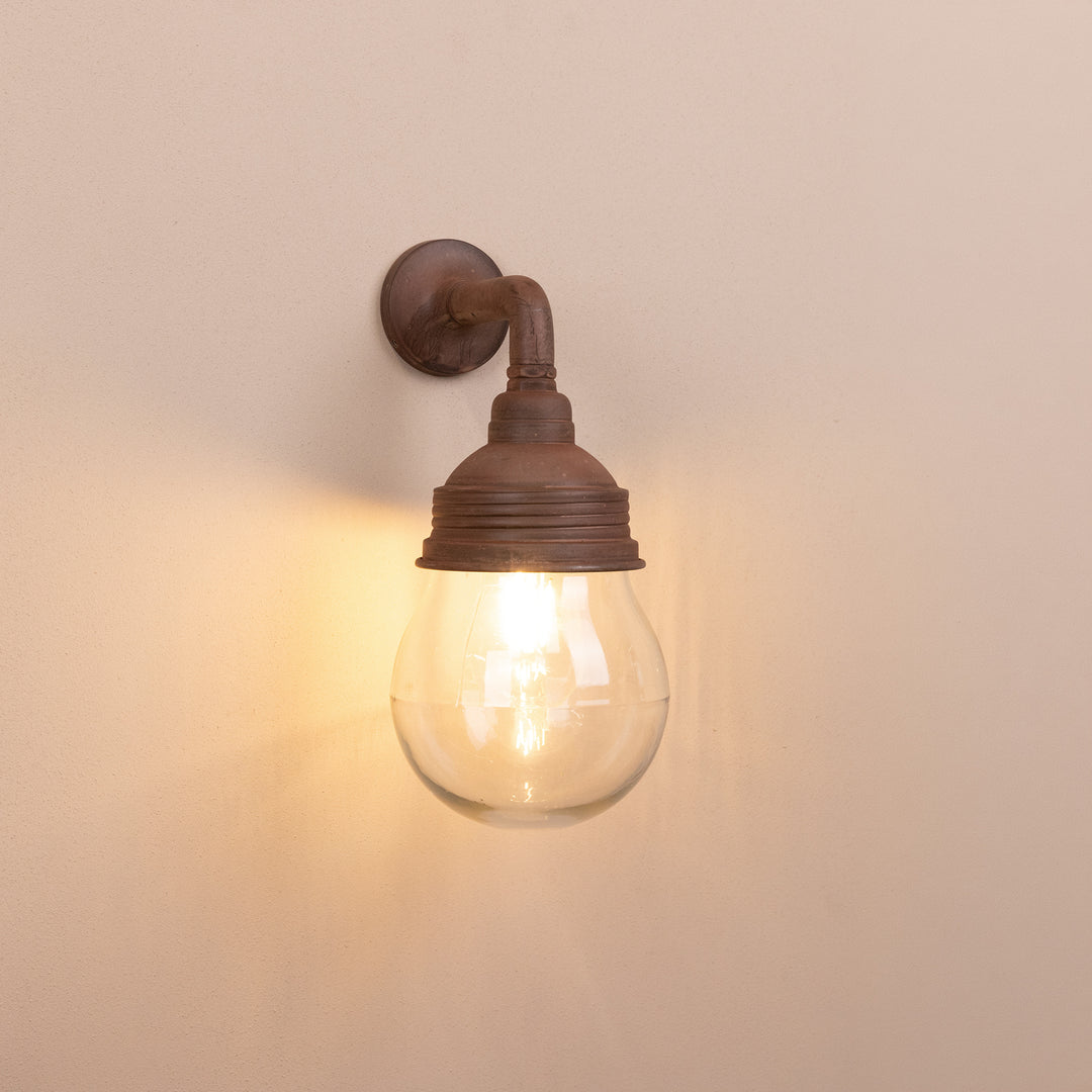 Front on image of the Agriframes Trowbridge Outdoor Wall Lamp in a Rust finish with its bulb illuminated, mounted on a neutral background. 