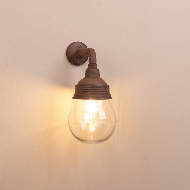 Front on image of the Agriframes Trowbridge Outdoor Wall Lamp in a Rust finish with its bulb illuminated, mounted on a neutral background. 