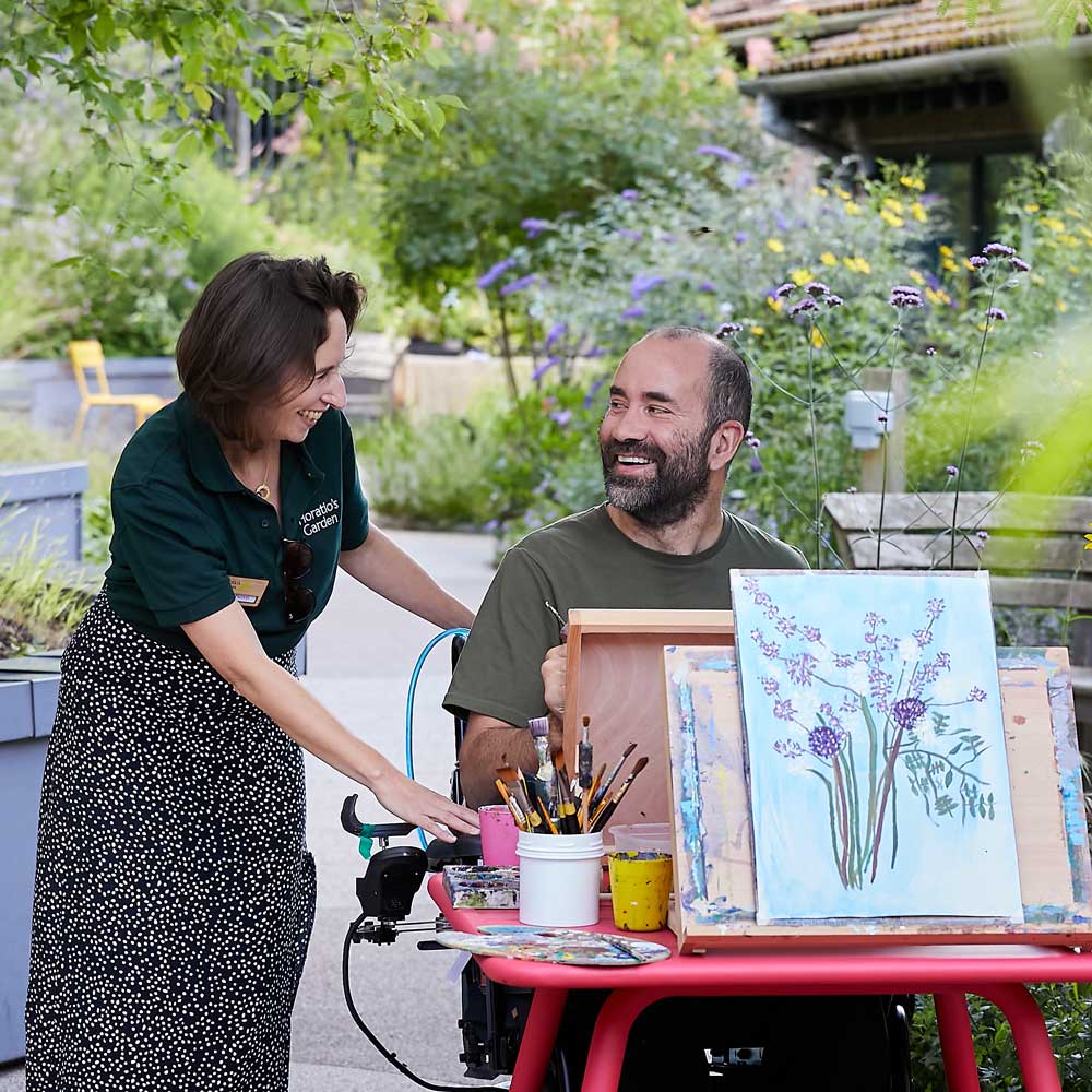 An arts workshop taking place in Horatio's Garden London and South East, a wheelchair bound man at an easel and a Horatio's Garden employe are smiling together.