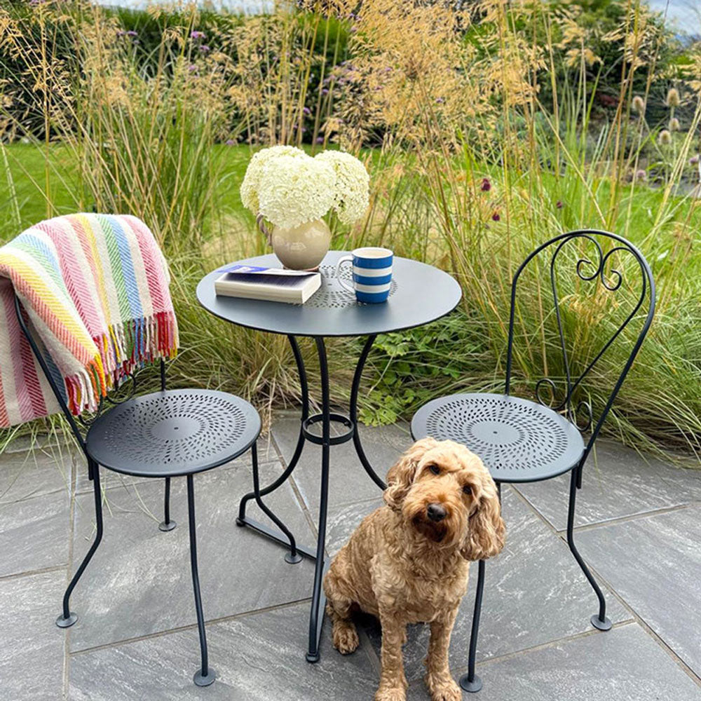 Outdoor setting with a small table and chairs, a dog sitting on the ground, and a colorful blanket draped over one of the chairs.