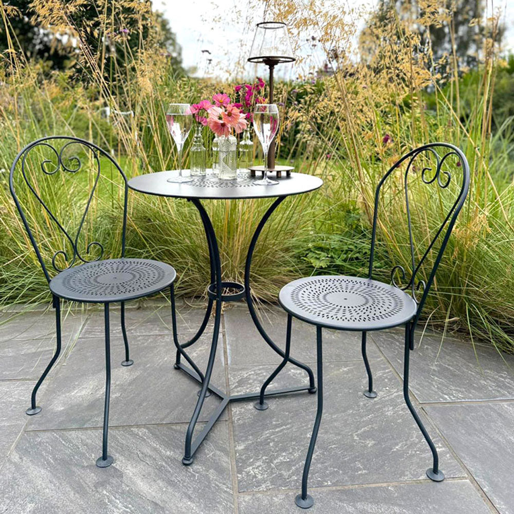 Black metal garden bistro set (table and chairs set) on a stone patio with flowers and glasses.