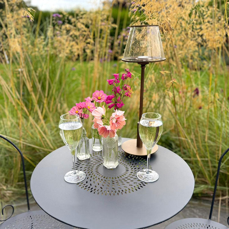 Outdoor setting with a bistro table featuring two glasses of champagne, a vase of flowers, and a tea light lamp, surrounded by tall grasses.