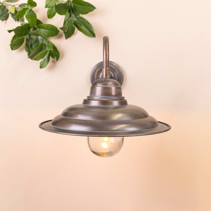 Copper wall-mounted lamp with illuminated bulb on a neutral background with decorative leaves in the top left corner.