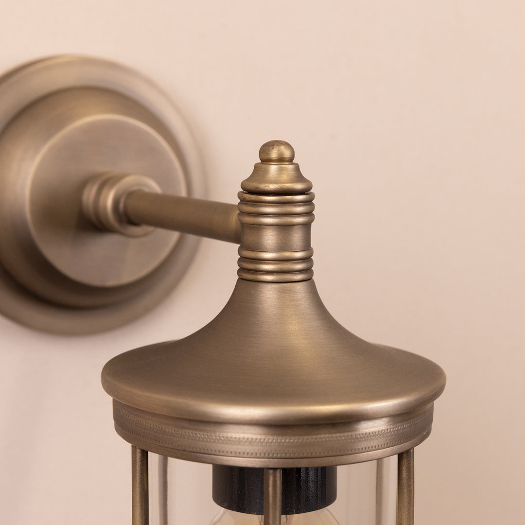 Closeup image of the base and stem of the Agriframes Hampstead Outdoor Wall Lamp in a Brass finish mounted on a neutral background. 