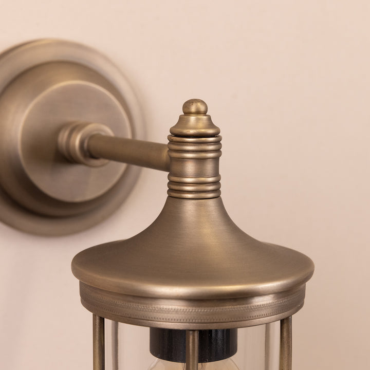Closeup image of the base and stem of the Agriframes Hampstead Outdoor Wall Lamp in a Brass finish mounted on a neutral background. 