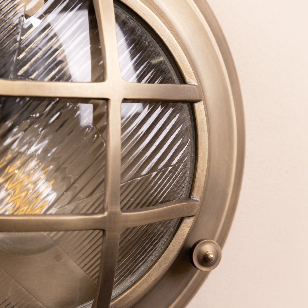 Closeup image of the Agriframes Looe Bulk Head Round Lantern in a Brass finish with its bulb switched off, mounted on a neutral background. 