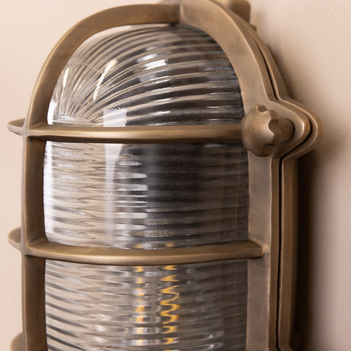 Closeup image of the Agriframes Bulk Head Oval Lantern in a Brass finish with its bulb switched off, mounted on a neutral background. 