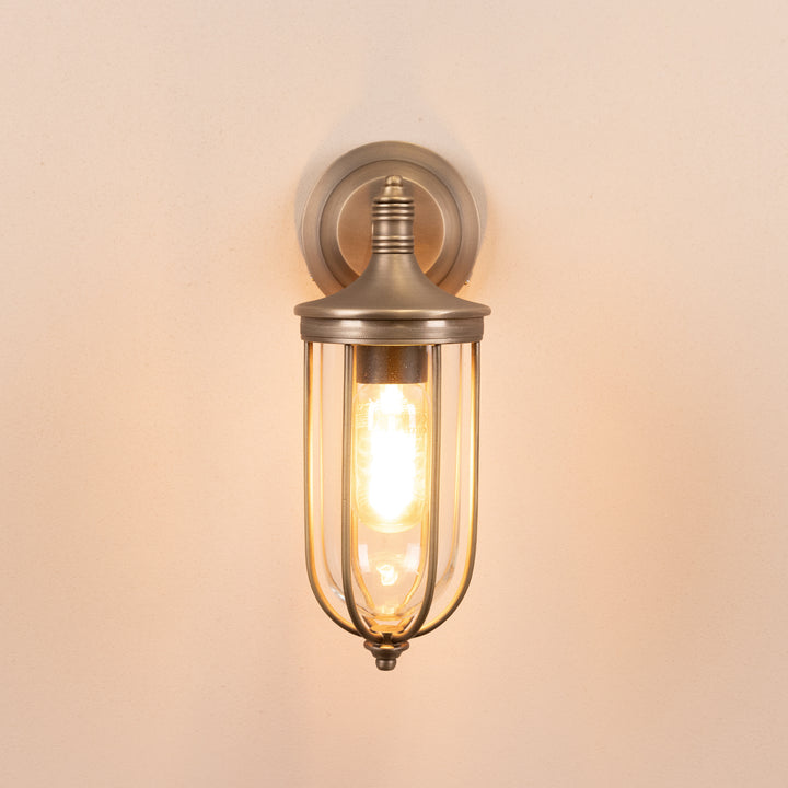 Front on image of the Agriframes Hampstead Outdoor Wall Lamp in a Brass finish with its bulb switched on, mounted on a neutral background.