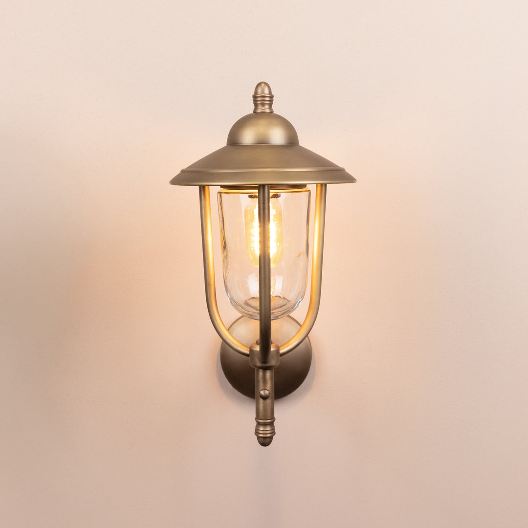 Front on image of the Agriframes Kensington Outdoor Wall Lamp in a Brass finish with its bulb switched on, mounted on a neutral background.