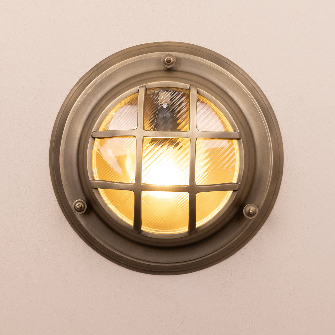 Front on image of the Agriframes Looe Bulk Head Round Lantern in a Brass finish with its bulb illuminated, mounted on a neutral background. 