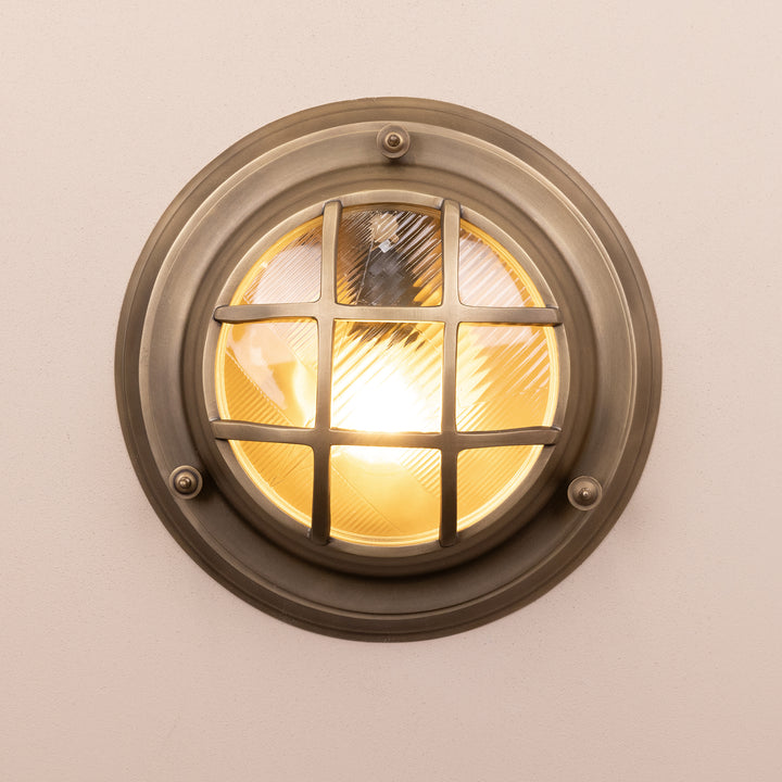 Front on image of the Agriframes Looe Bulk Head Round Lantern in a Brass finish with its bulb illuminated, mounted on a neutral background. 