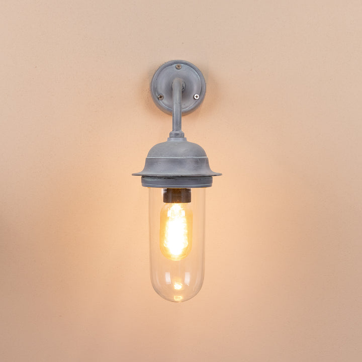 Front on image of the Agriframes Pewsey Outdoor Wall Lamp in an Old Iron finish with its bulb illuminated, mounted on a neutral background. 