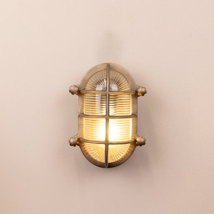 Front on image of the Agriframes Bulk Head Oval Lantern in a Brass finish with its bulb illuminated, mounted on a neutral background. 