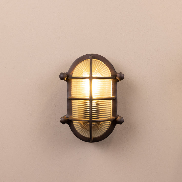 Front on image of the Agriframes Salcombe Bulk Head Oval Lantern in a Bronze finish with its bulb illuminated, mounted on a neutral background. 