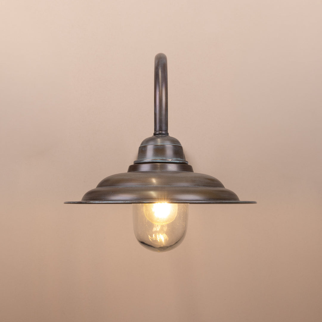 Front on image of the Agriframes Tetbury Outdoor Wall Lamp in a Copper Patina finish with its bulb illuminated, mounted on a neutral background. 