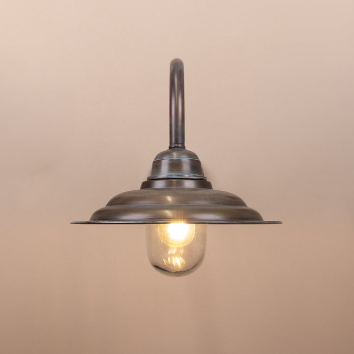 Front on image of the Agriframes Tetbury Outdoor Wall Lamp in a Copper Patina finish with its bulb illuminated, mounted on a neutral background. 